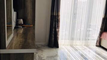 Una habitación con piso de mármol, un mueble de madera, un televisor de pantalla plana, una cafetera y una ventana con cortinas traslúcidas.