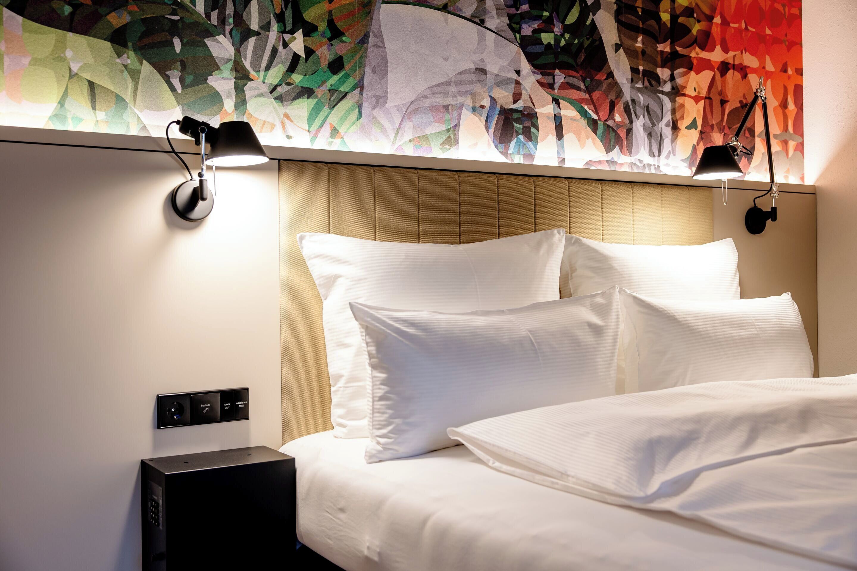 A hotel room with a bed, two bedside lamps, a control panel, and a colorful abstract wall art.