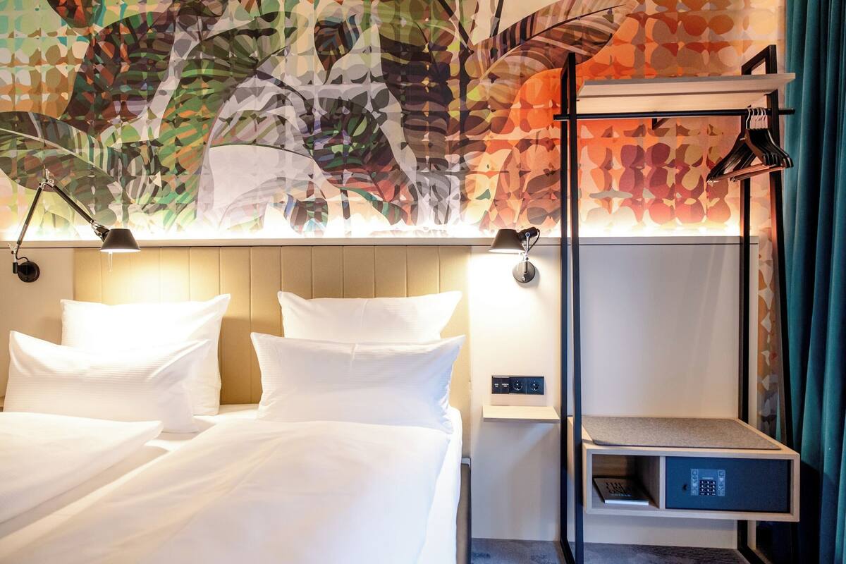A hotel room with a bed, bedside lamps, a nightstand with a lamp, a small shelf, and a decorative wall with leaf patterns.