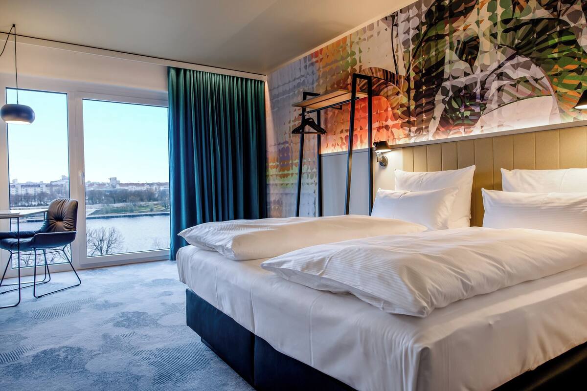 A hotel room with a large bed, a desk with a chair, a view of the city through a large window, and a colorful abstract wall mural.
