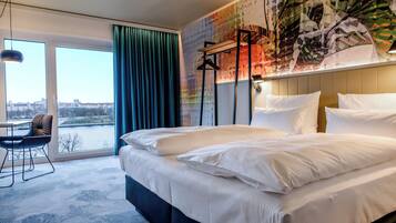 A hotel room with a large bed, a desk with a chair, a view of the city through a large window, and a colorful abstract wall mural.