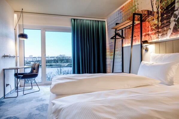 Une chambre d’hôtel avec un grand lit, un bureau avec une chaise, une vue sur la ville par la fenêtre et une fresque murale aux couleurs vives.