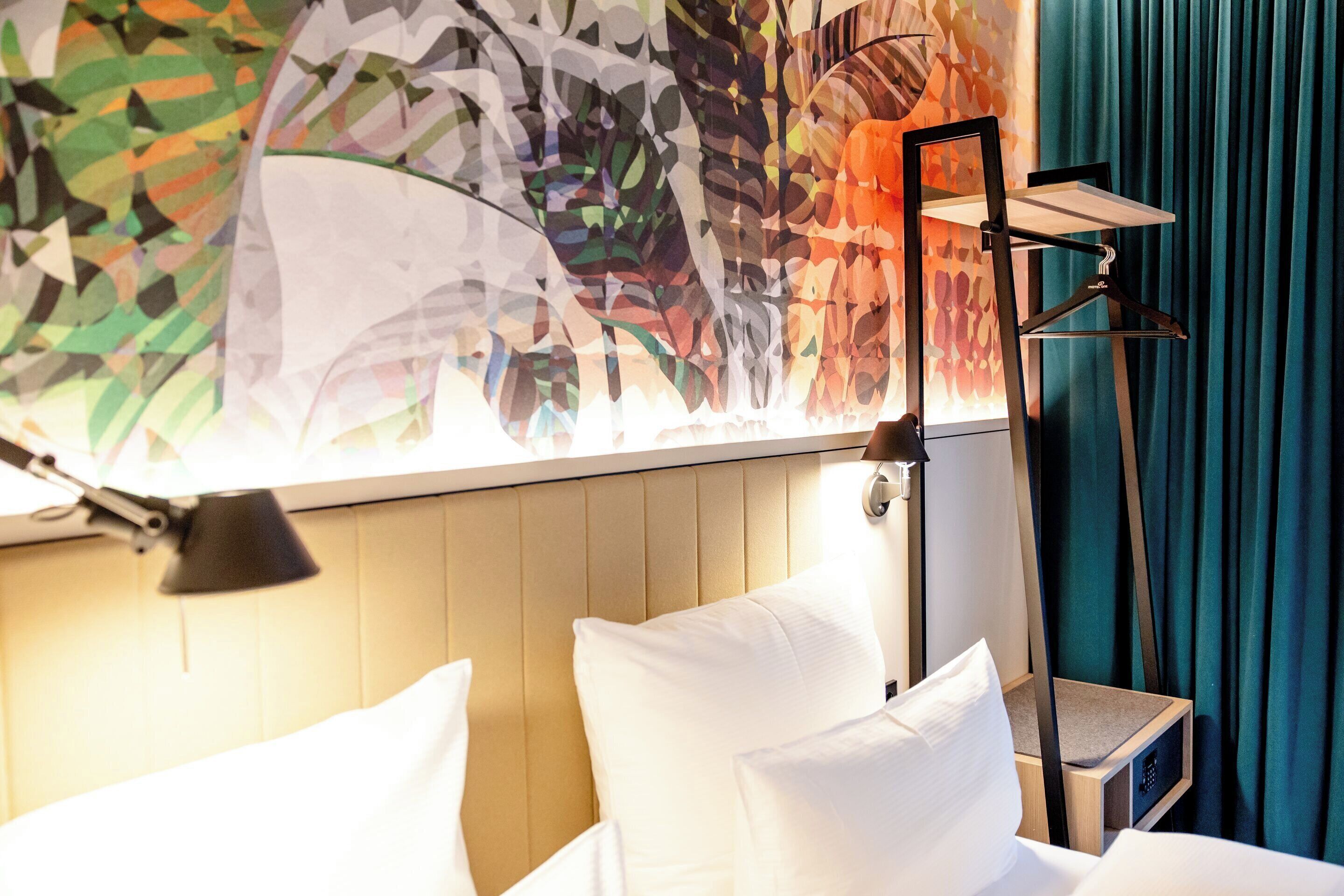 Una habitación de hotel con una cama, una mesita de noche, una lámpara y un cuadro abstracto de colores en la pared.