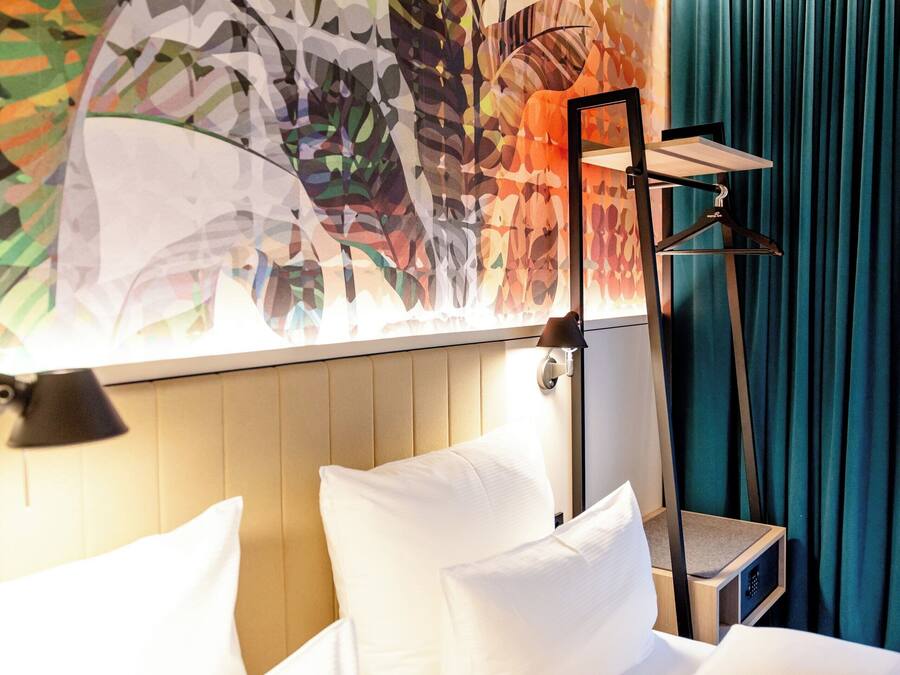 Una habitación de hotel con una cama, una mesita de noche, una lámpara y un cuadro abstracto de colores en la pared.
