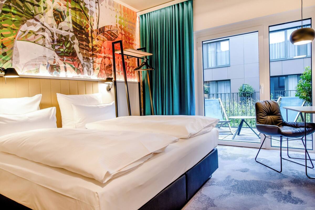 A hotel room with a large bed, a chair, a table, and a view of the outside balcony.