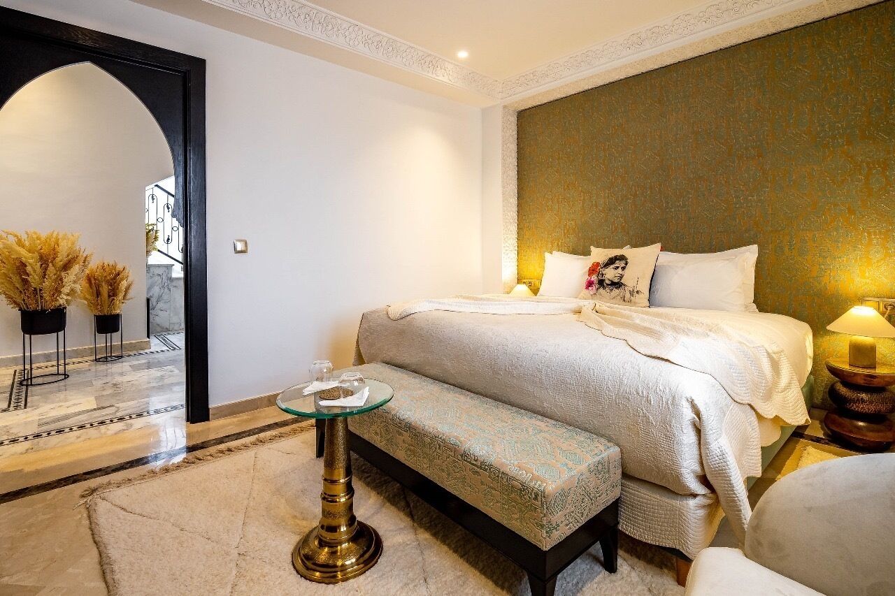 A hotel room with a large bed, a bench, a small table, and a decorative mirror.