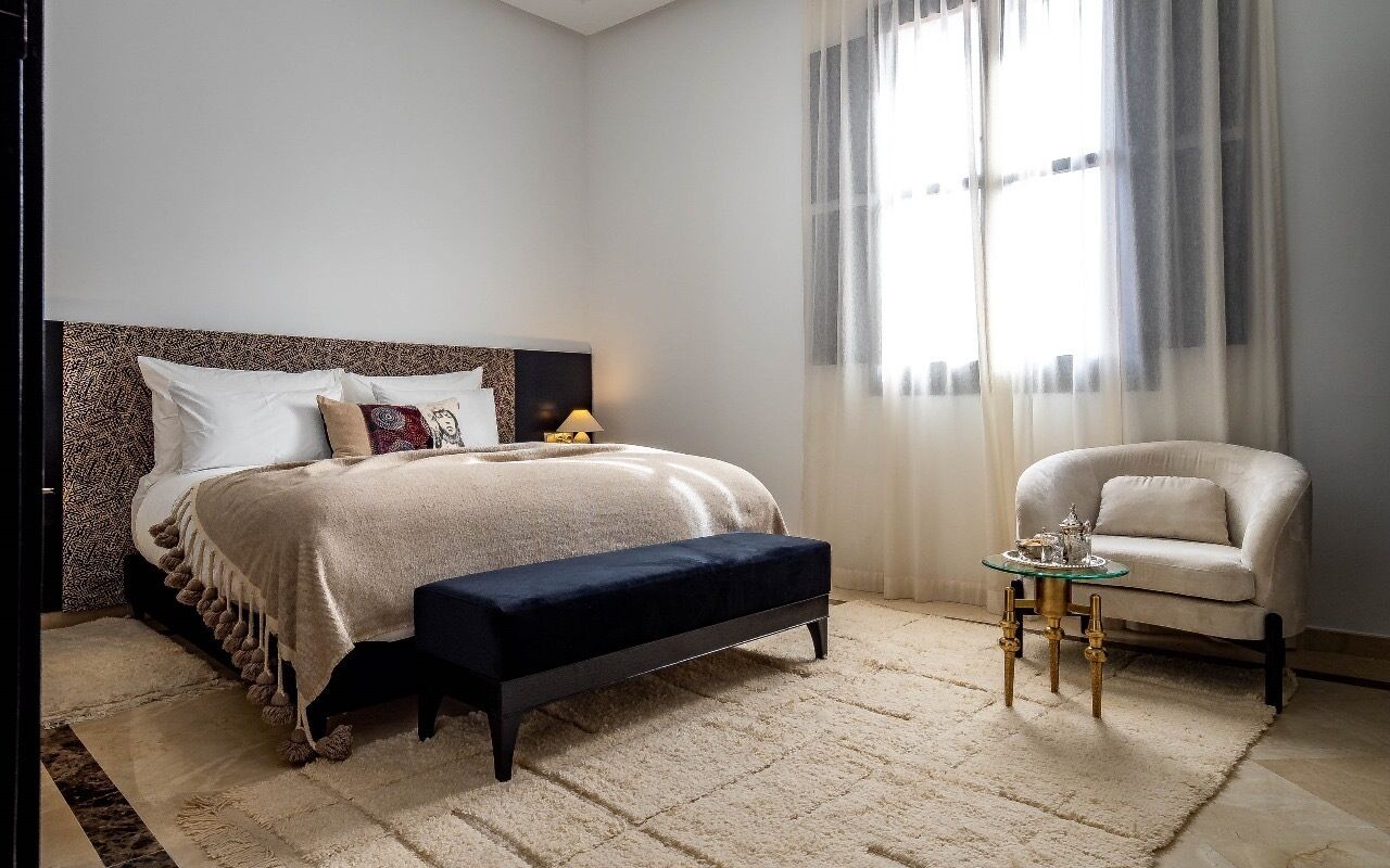 Un dormitorio con cama, banco, silla y una mesita de noche con lámpara.