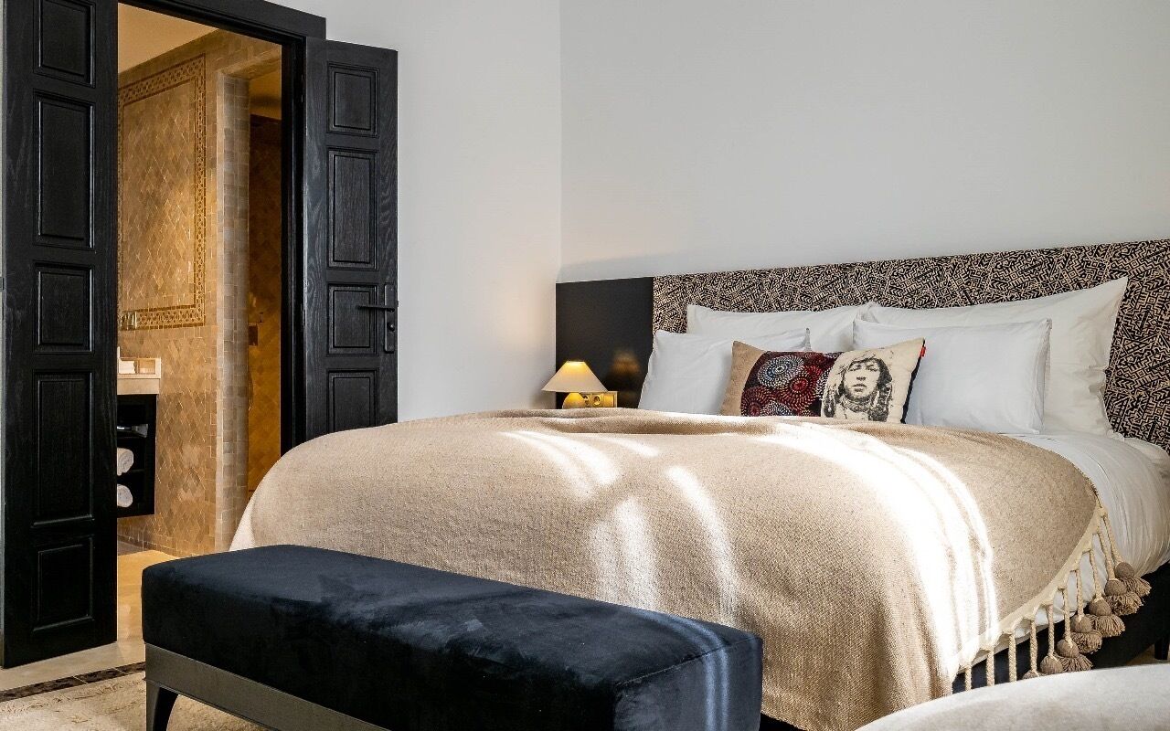 A bedroom with a bed, a bedside table with a lamp, a bench, and a door leading to a bathroom.