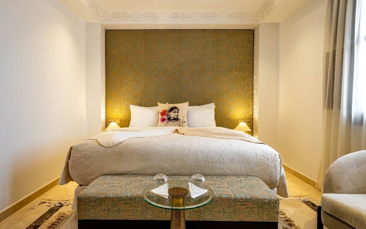 A neatly arranged bedroom with a large bed, a bedside table with two glasses, and a patterned ottoman.
