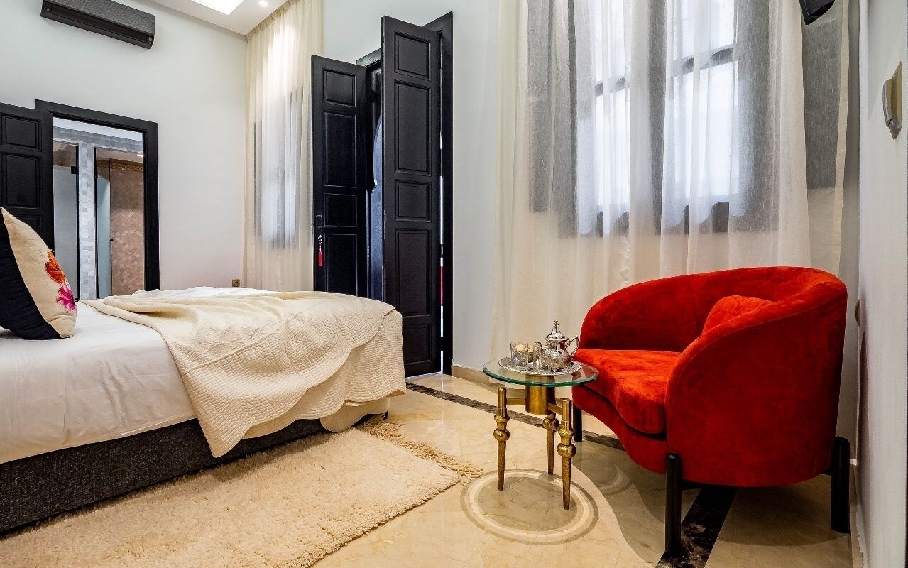 Une chambre à coucher avec un lit, un fauteuil rouge, une petite table avec un plateau, et une grande fenêtre avec des rideaux légers.