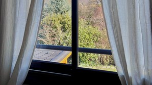 A window with sheer white curtains, revealing a mountain landscape with snow-capped peaks and dense forest.
