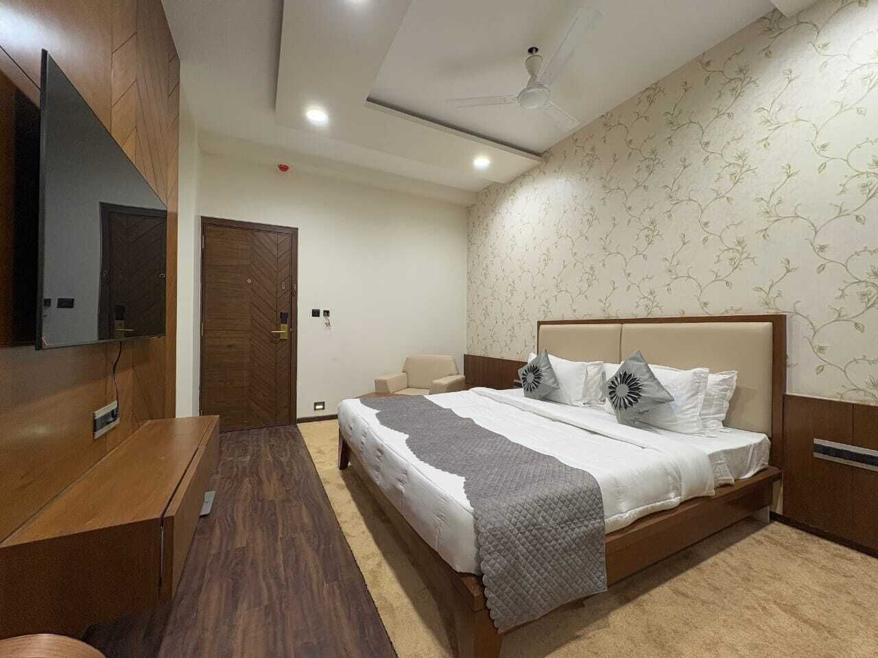 A bedroom with a bed, a TV mounted on the wall, a wooden wardrobe, and a ceiling fan.