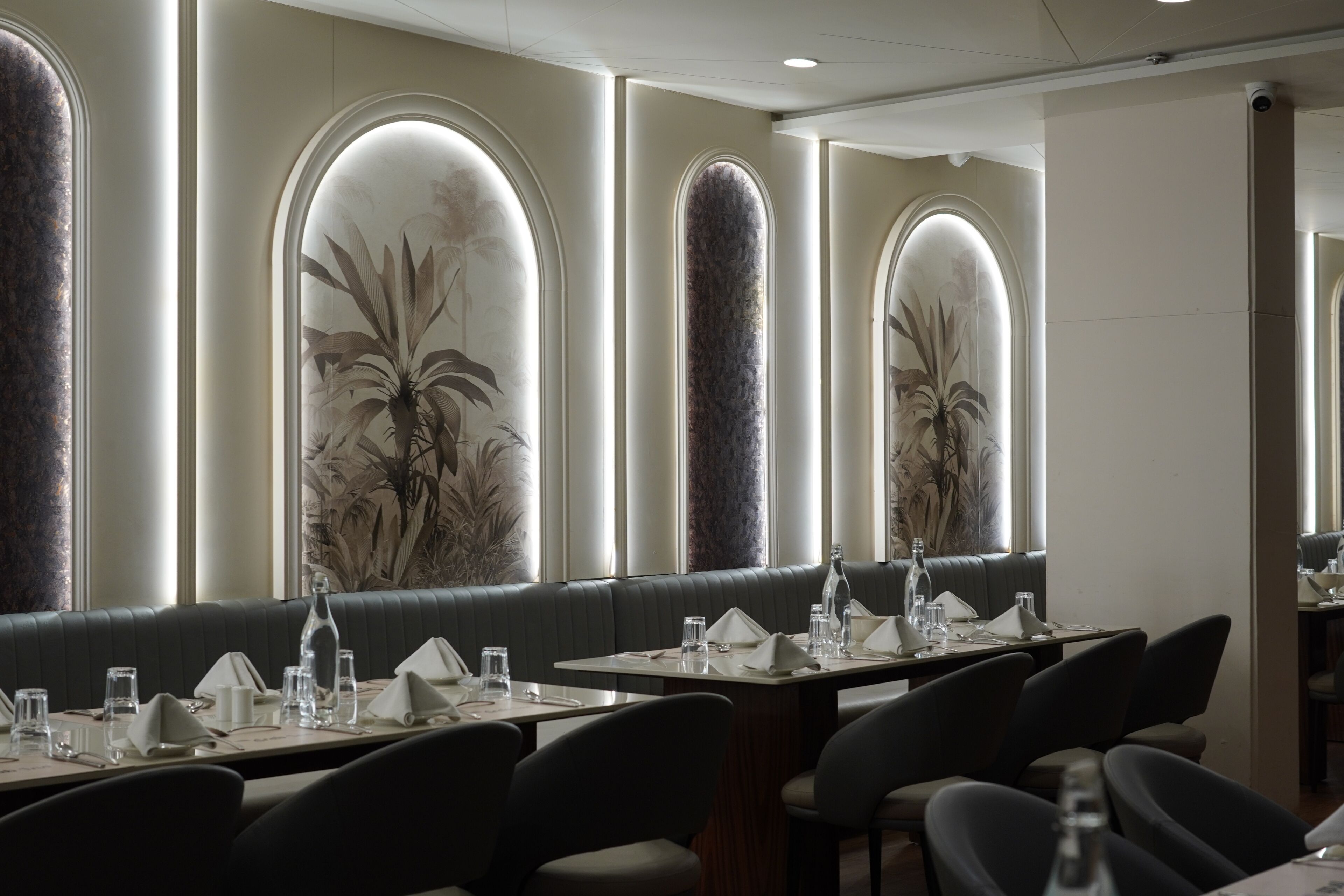 Un restaurante con sillas de respaldo alto, mesas dispuestas con vasos y cubiertos, y amplias ventanas en arco decoradas con murales de plantas.