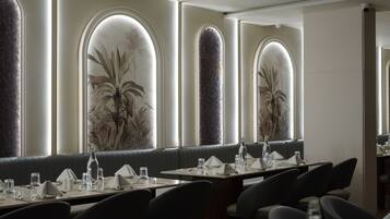 Un restaurante con sillas de respaldo alto, mesas dispuestas con vasos y cubiertos, y amplias ventanas en arco decoradas con murales de plantas.