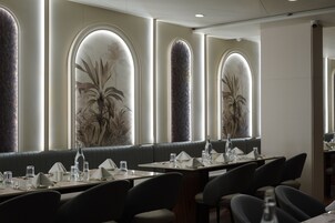 Un restaurante con sillas de respaldo alto, mesas dispuestas con vasos y cubiertos, y amplias ventanas en arco decoradas con murales de plantas.