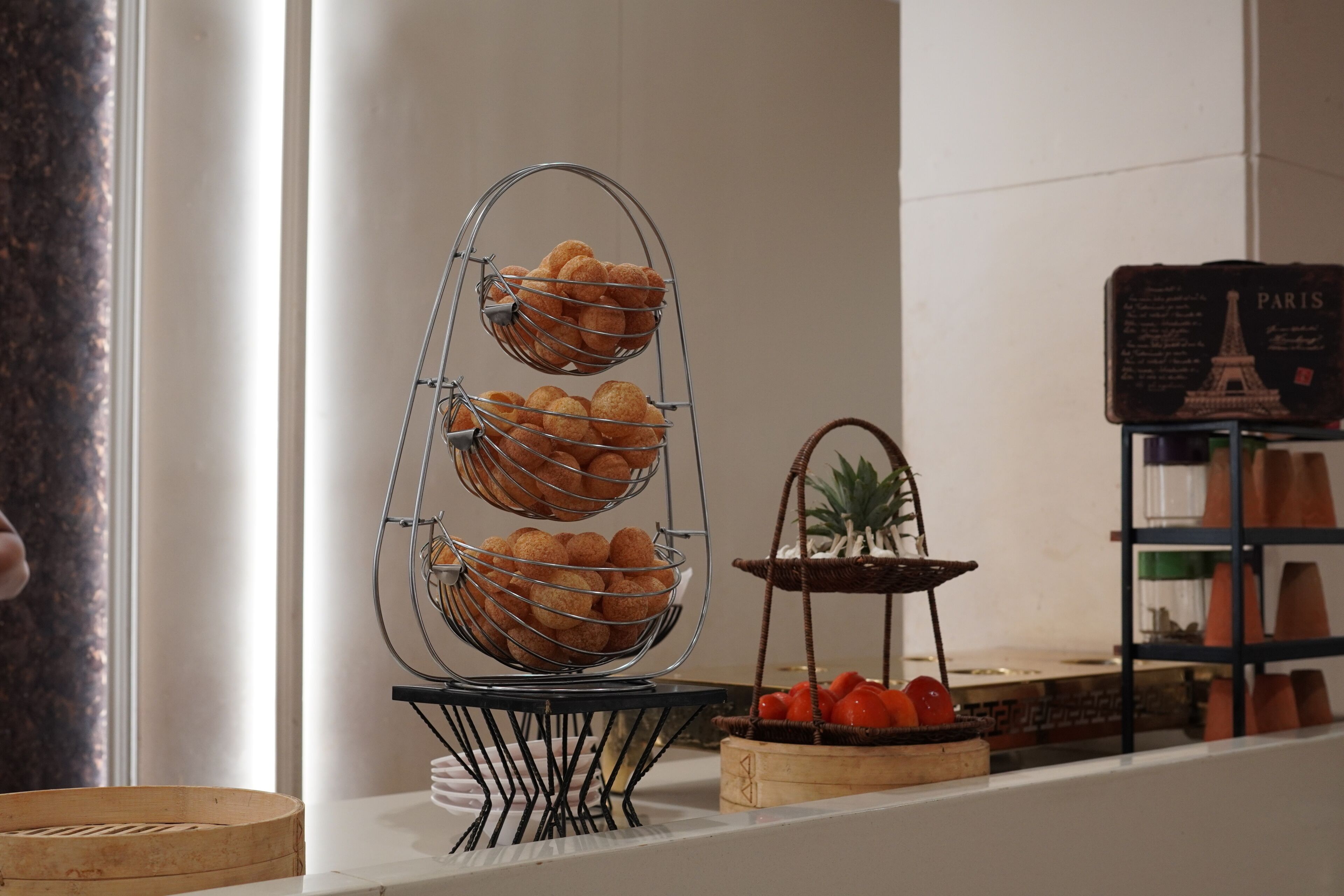 A buffet setup with a tiered metal rack holding bread, a wicker basket with tomatoes, and a sign with 'PARIS' and an Eiffel Tower illustration.