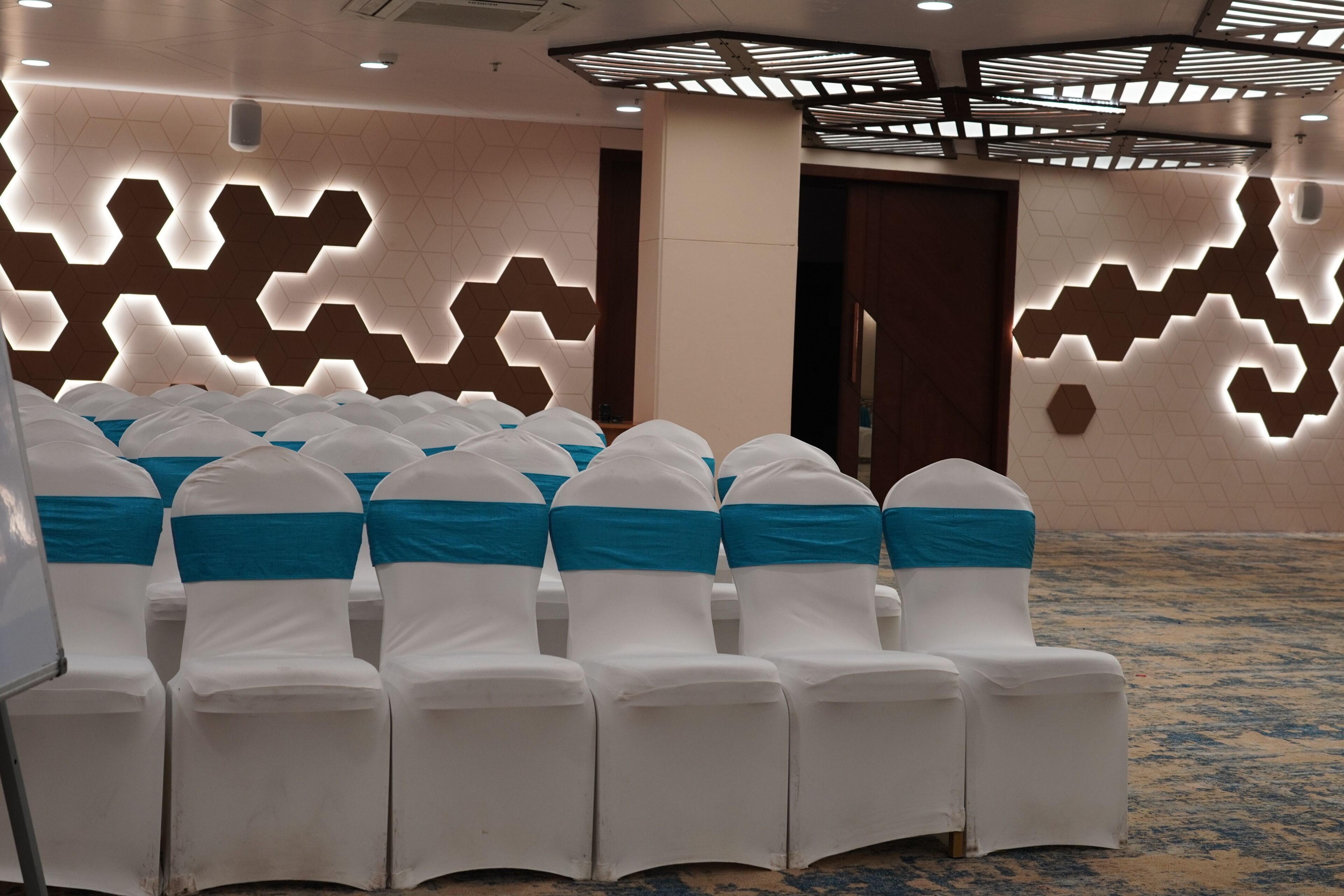 Rows of chairs with white covers and blue sashes, arranged in a conference room with hexagonal light patterns on the walls.