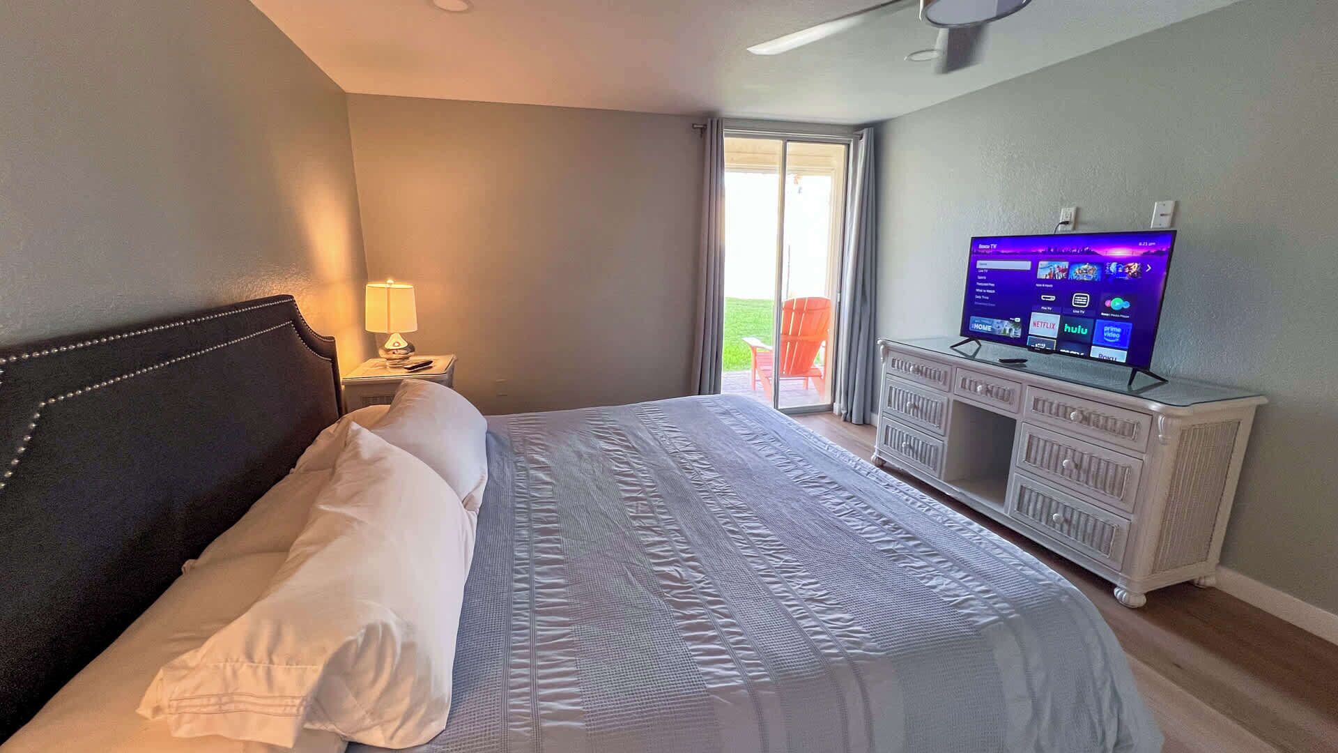 A hotel room with a bed, a TV, a nightstand, and a view of the outdoors.