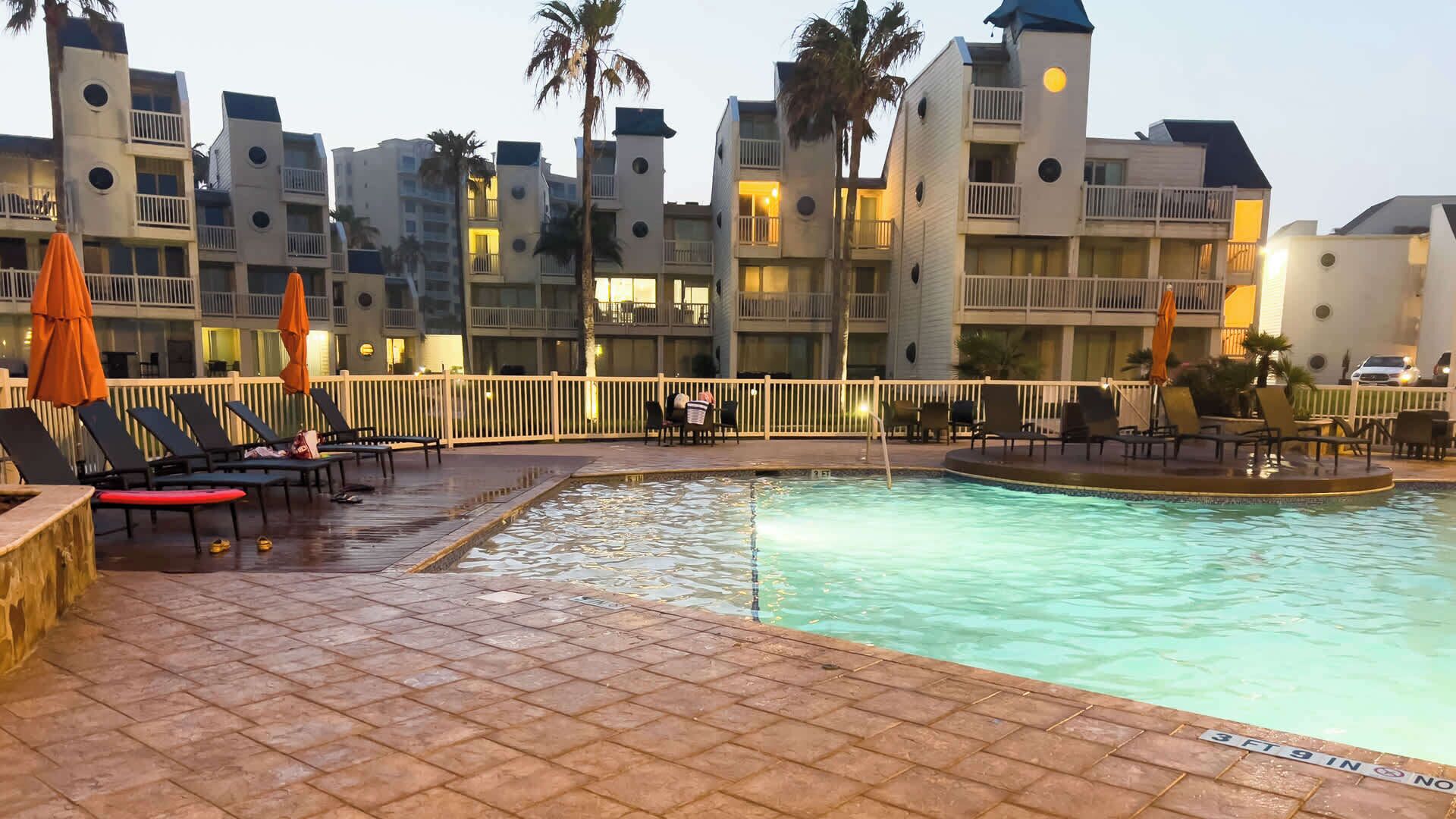 A hotel pool area with sun loungers and umbrellas, surrounded by multi-story buildings with balconies.