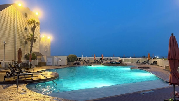 Area piscina con lettini e ombrelloni, circondata da una recinzione bianca, e un edificio con numerose finestre e luci.