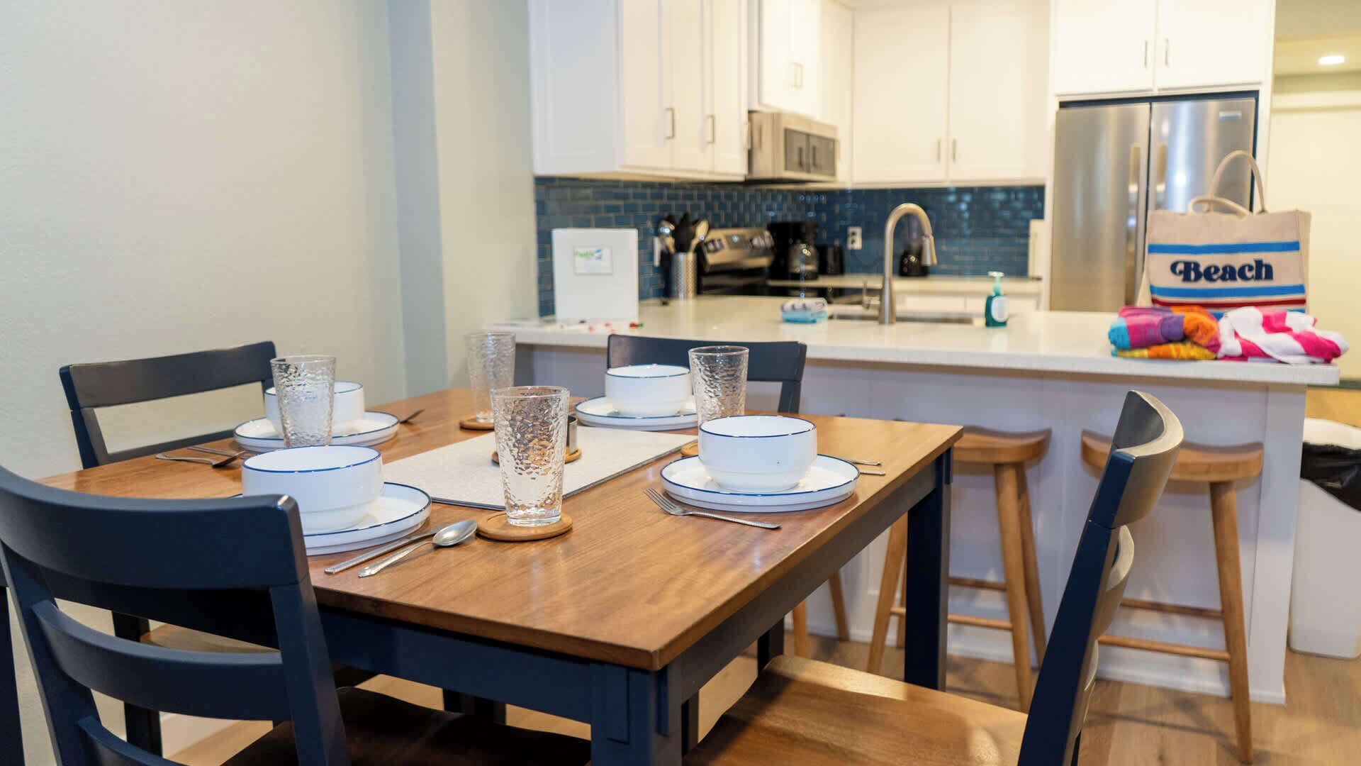 A dining area with a wooden table set for four, surrounded by black chairs. A kitchen with white cabinets and a stainless steel refrigerator is visible in the background.