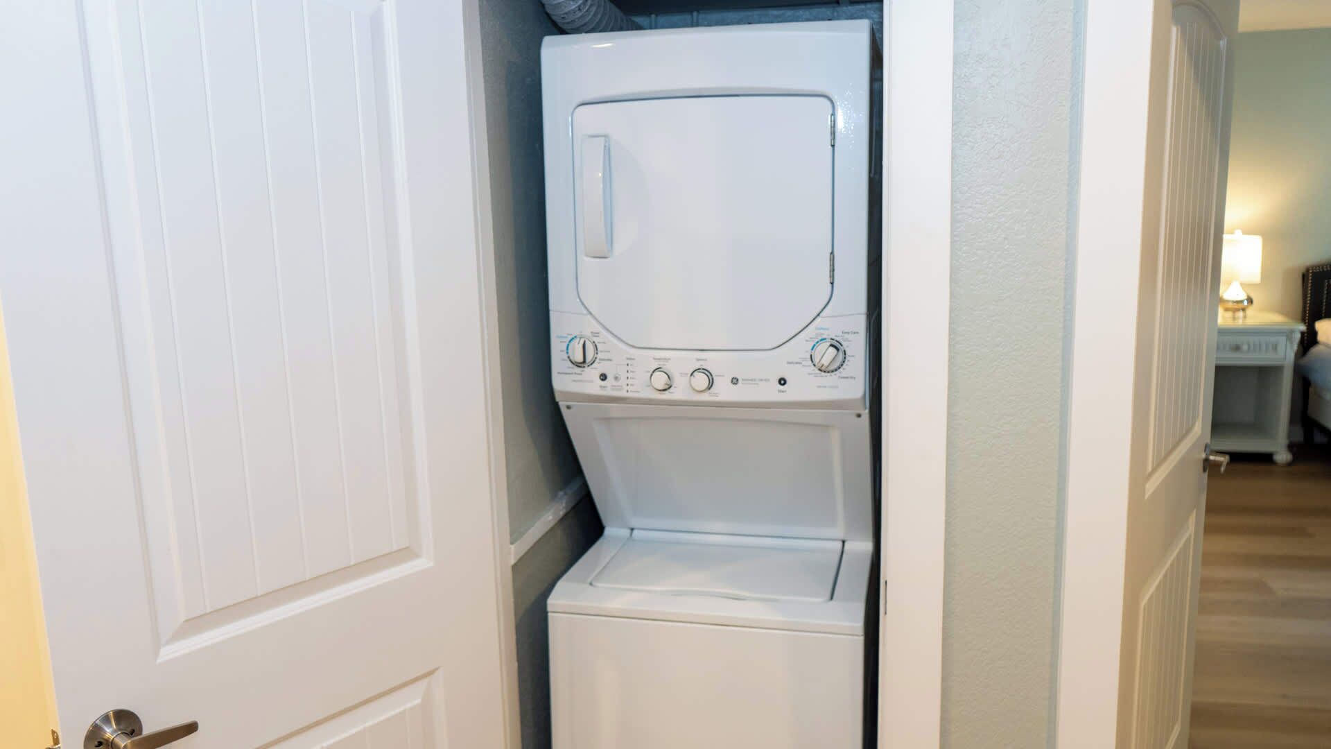 A compact, white laundry room with a built-in washer and dryer, a door, and a small nightstand with a lamp in the background.