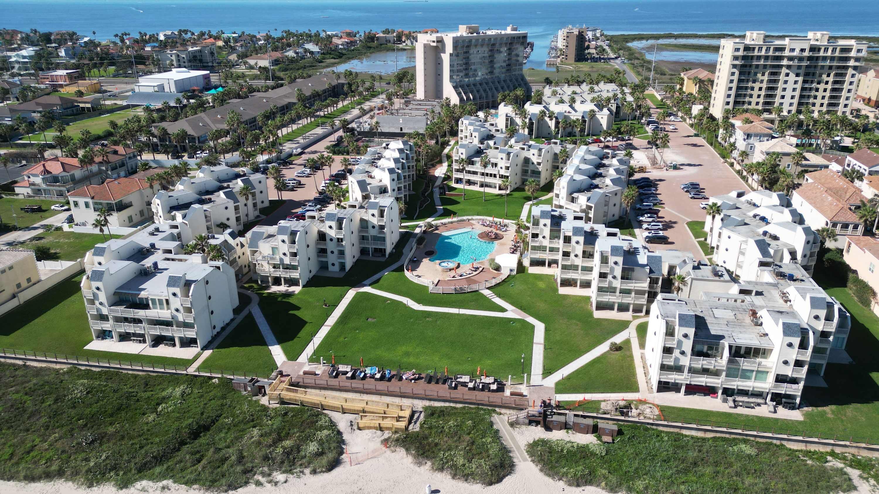 Vue aérienne d’un complexe hôtelier comprenant plusieurs bâtiments blancs, un espace piscine central et un accès à la plage.