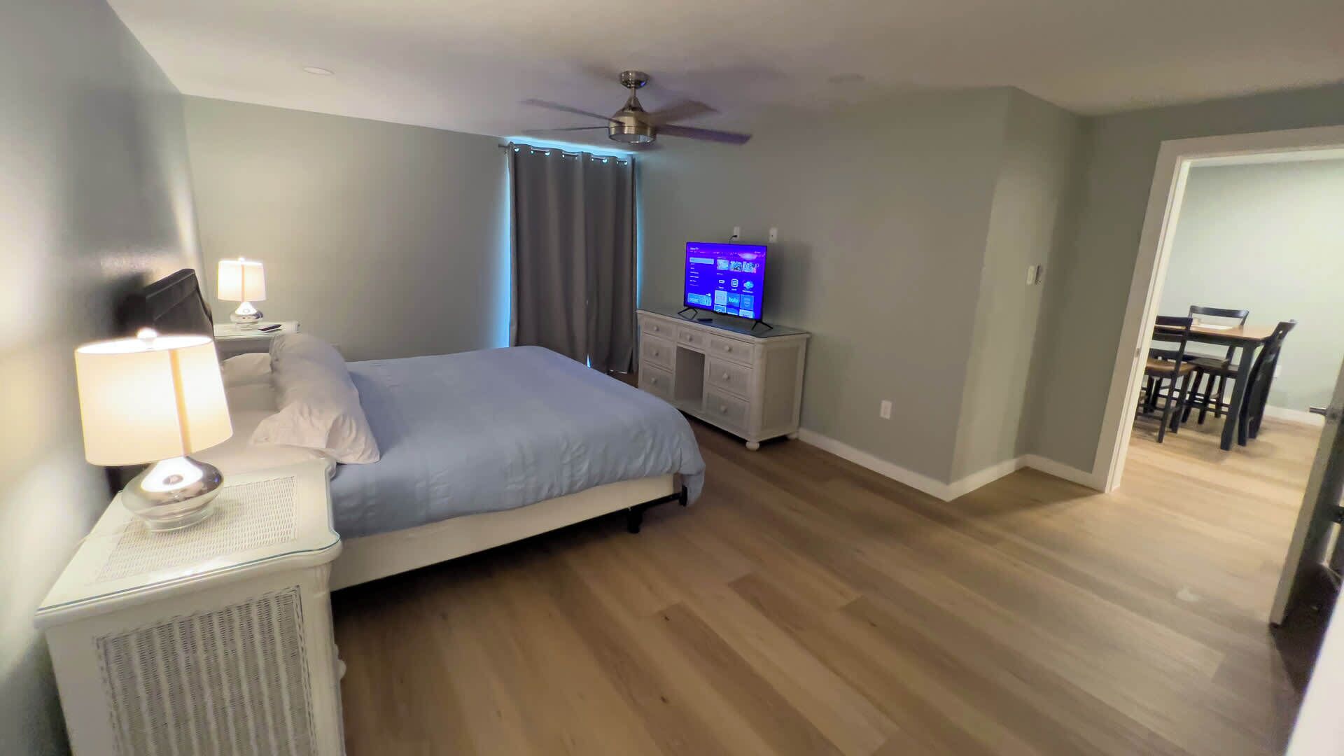 A bedroom with a bed, a desk with a computer, a ceiling fan, and a dining area with chairs.