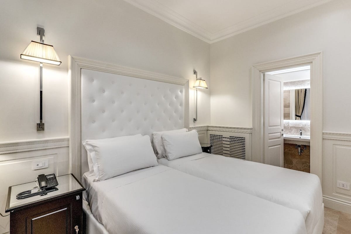 Habitación de hotel con una cama grande, dos lámparas de noche, un teléfono en la mesita y un baño visible a través de una puerta abierta.