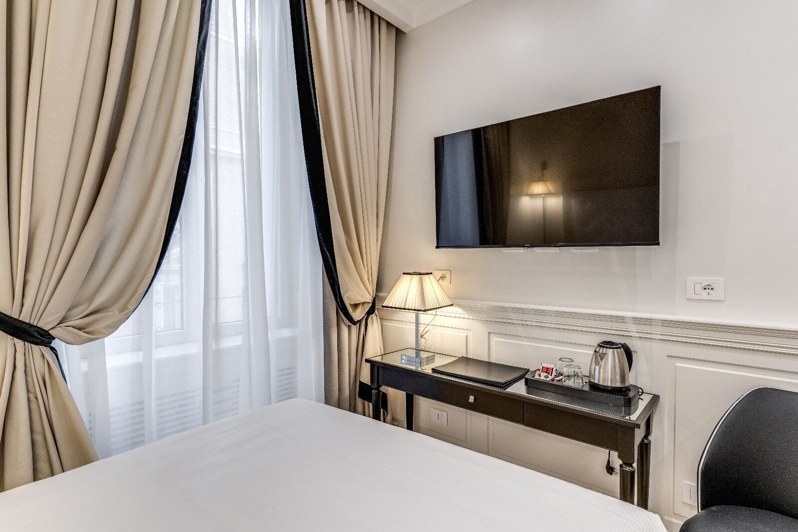 Quarto de hotel com cama, escrivaninha com luminária, televisão e cadeira.