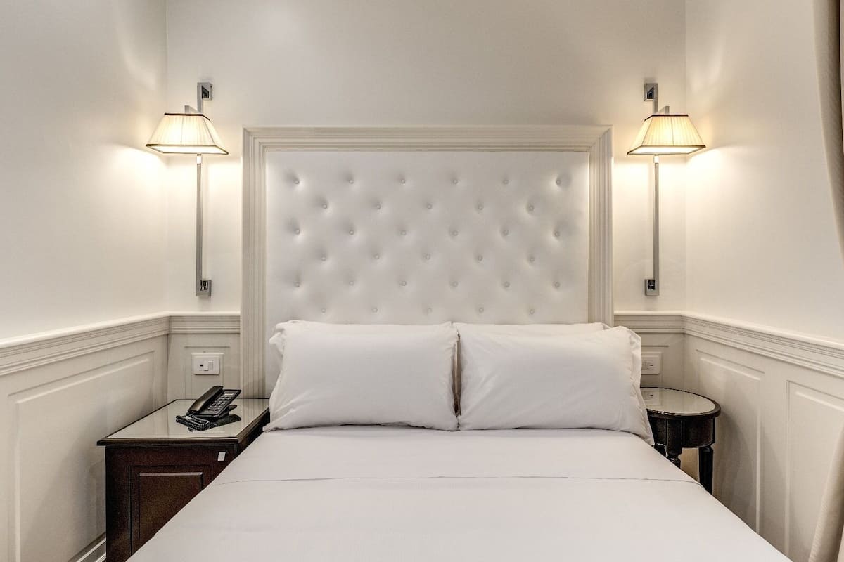 Una cama bien hecha con sábanas blancas y dos almohadas, flanqueada por dos mesitas de noche con lámparas, frente a una pared blanca con cabecera capitonada.