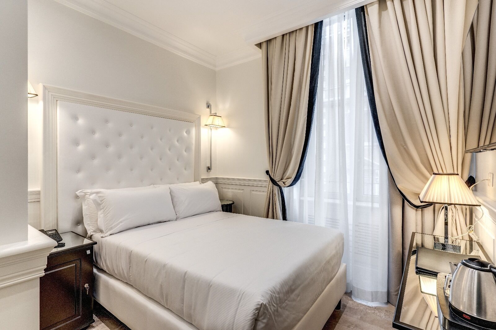 Camera d'albergo ben ordinata con un letto grande, un comodino, una lampada e vista all'esterno attraverso tende leggere.