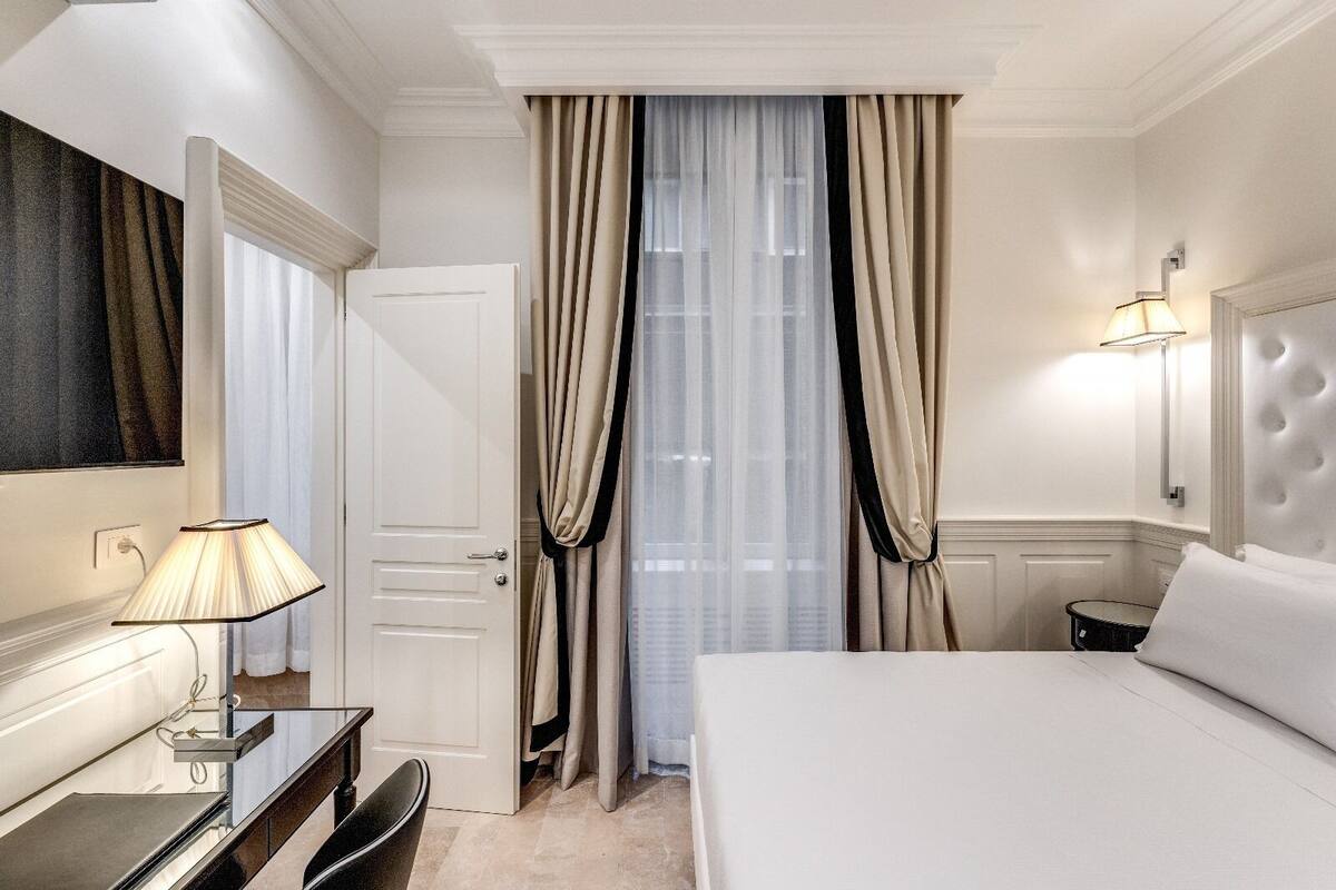 Habitación de hotel con cama, escritorio con lámpara, espejo, ventana con cortinas e iluminación en la pared.
