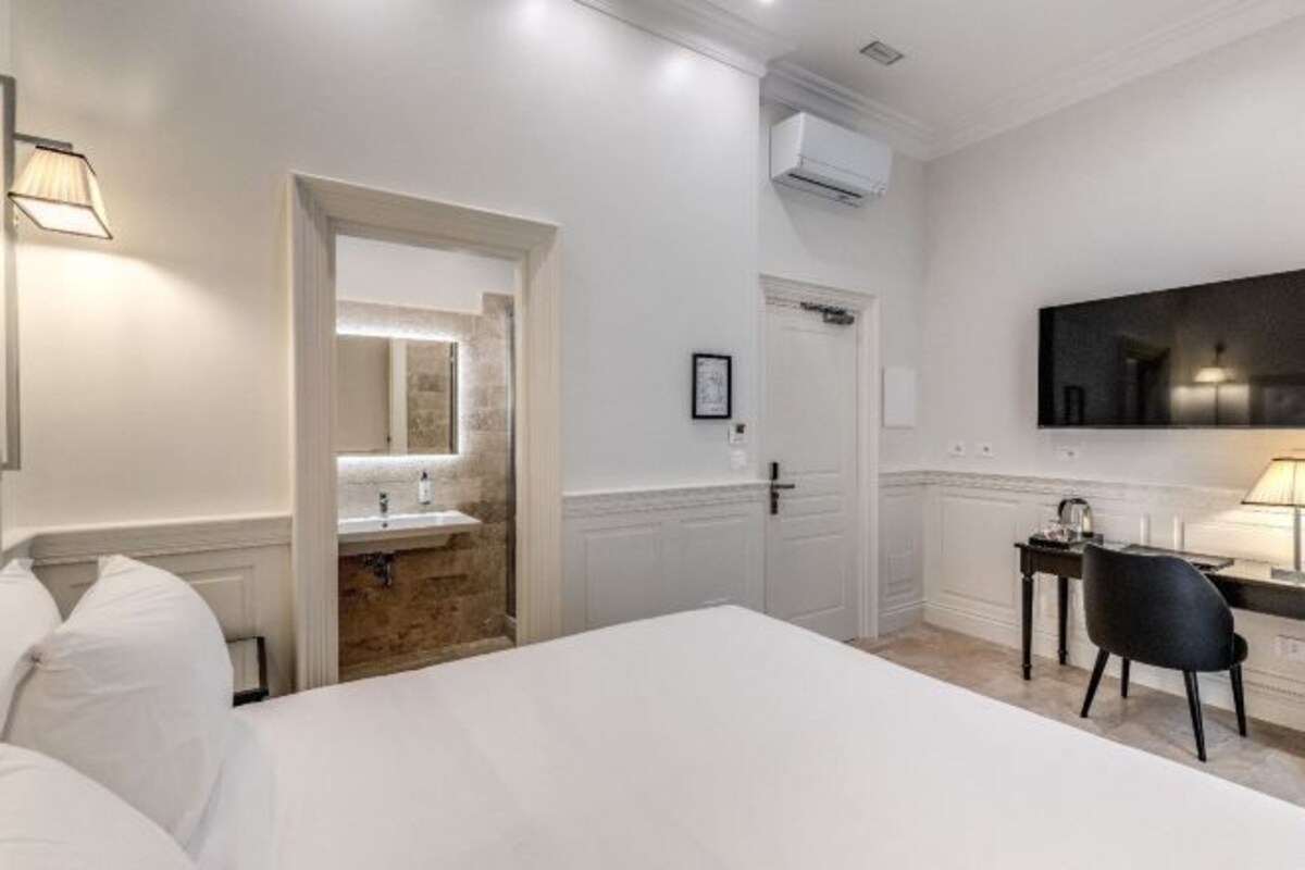 A hotel room with a large bed, a desk with a chair, a TV mounted on the wall, and a bathroom with a mirror and sink.