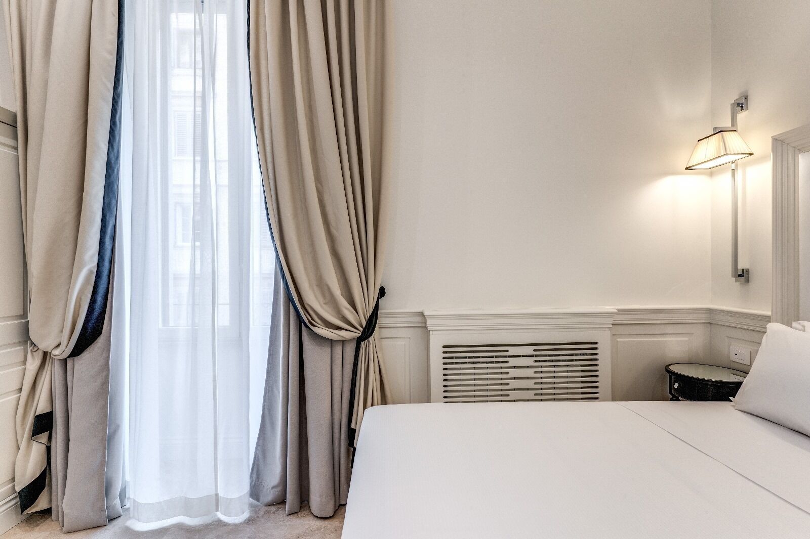 A hotel room with a bed, a window with sheer curtains, a wall-mounted lamp, and a radiator.