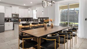 Una cocina moderna con armarios blancos, una isla central con asientos, y un comedor con una mesa grande y sillas.