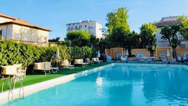 A swimming pool with lounge chairs and a grassy area, surrounded by buildings and trees. - HOTEL YOUNG PEOPLE UNDER 40 (Rimini)