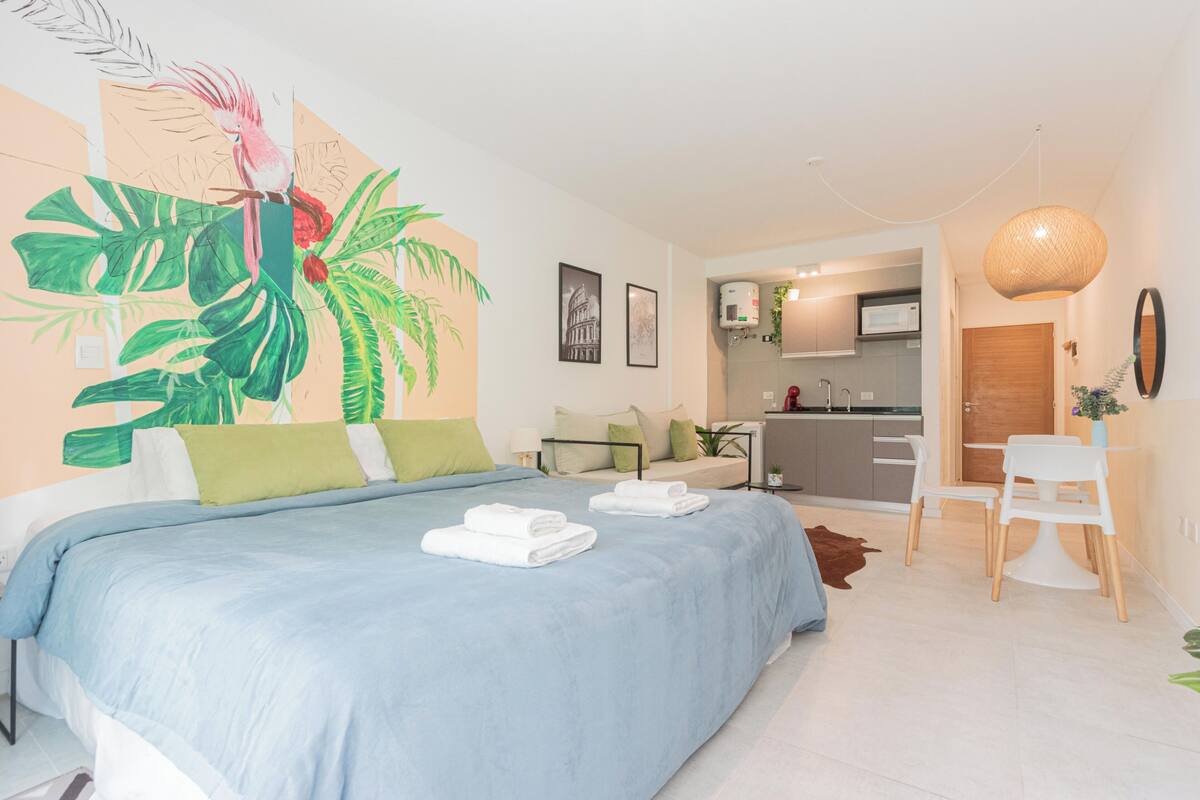 A modern hotel room with a large bed, a kitchenette, and a dining area.