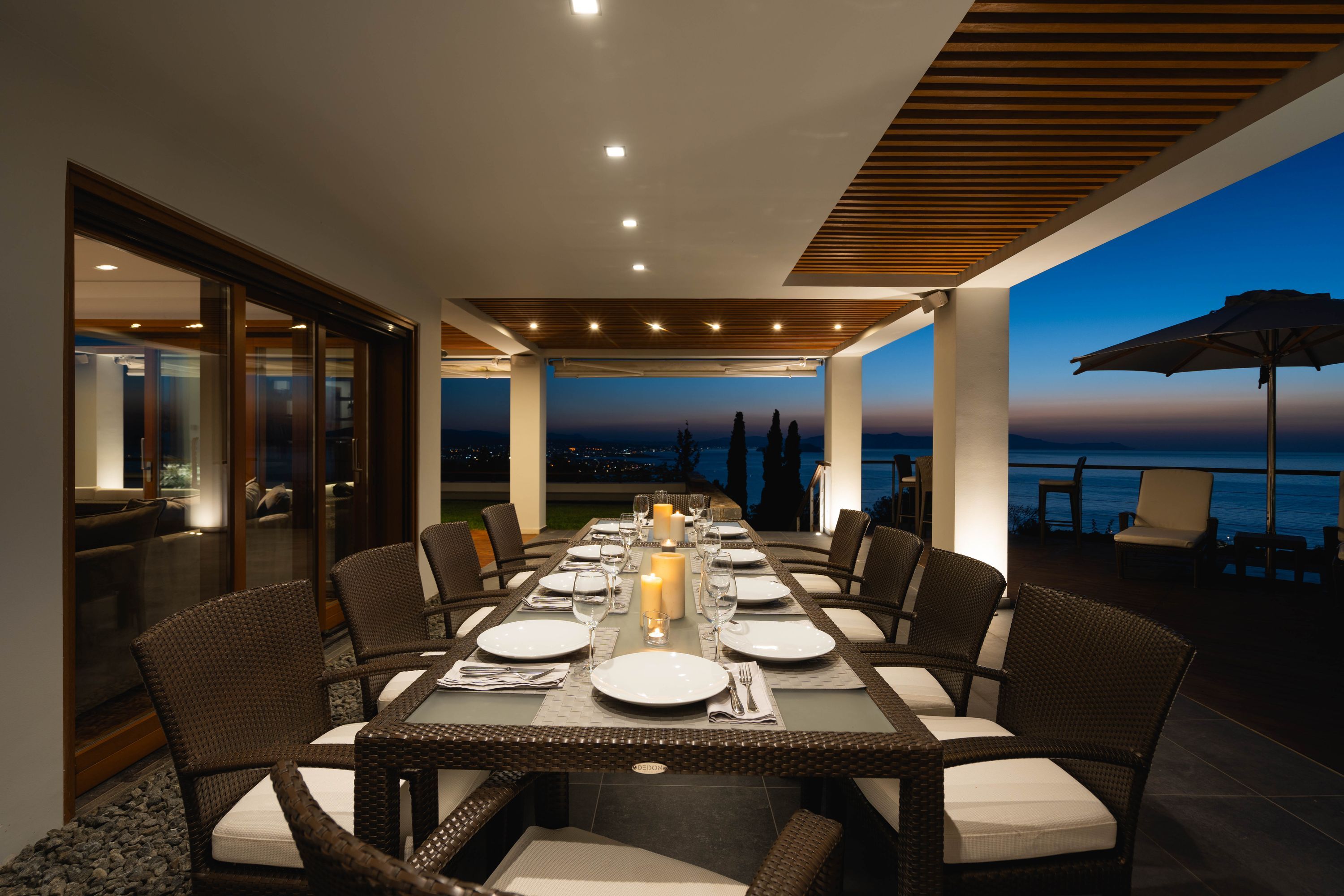 An outdoor dining area with a long table set for a meal, surrounded by wicker chairs, and overlooking a scenic view.