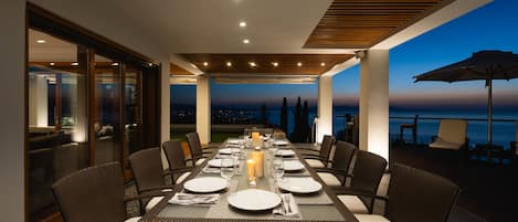 Un área de comedor al aire libre con una larga mesa lista para una comida, rodeada de sillas de mimbre y con vistas a un paisaje pintoresco.