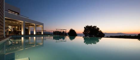 A modern infinity pool with clear water reflecting the sky, a covered outdoor area with seating, and a view of the sunset over the water.