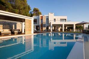 A modern poolside area with a clear pool, lounge chairs, and a multi-story building with large windows and outdoor seating. - Villa Terra Creta Luxury Retreat and Spa (Chania)