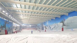 An indoor ski slope with artificial snow, ski lifts, and a mural of a snowy mountain.