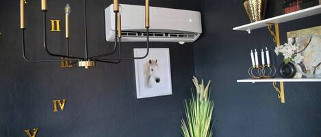 Une pièce avec un mur sombre, des lettres dorées, un lustre, un climatiseur, un tableau encadré, un vase avec des plantes et une étagère avec des bougies et des livres.
