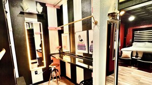 A hotel room with a bed, a vanity with a mirror, a stool, and a wall with framed items. - Farmhouse outskirts of Norman’s “Okla. University” fastWiFi, 2-75”TVs, KING BED (Noble)