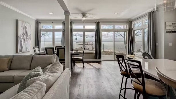 Una sala de estar moderna con un sofá, una mesa de comedor y sillas, ventanas grandes con vistas, y un ventilador de techo.