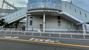 Un bâtiment moderne doté d’une façade en verre incurvée, d’une entrée imposante avec une rampe d’accès et d’une zone délimitée par une clôture à l’avant.