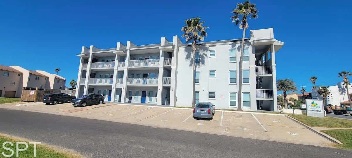 Un hotel de varios pisos con fachada blanca, balcones y un estacionamiento con varios coches.
