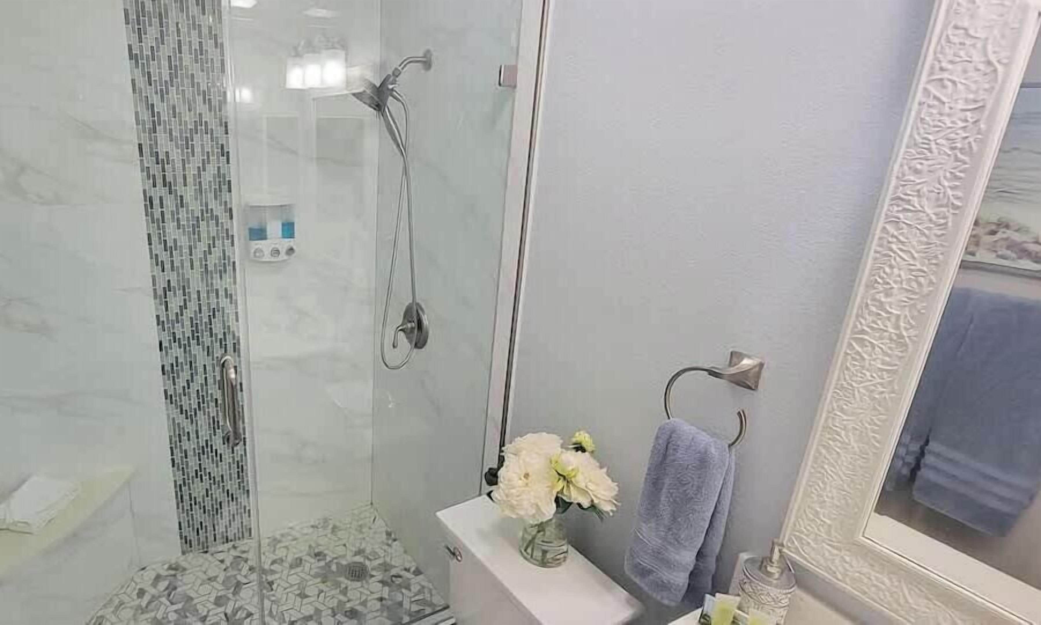 Et badeværelse med et glasafskærmet brusebad, en hvid vask med en vase blomster og et blåt håndklæde hængende på en bøjle.