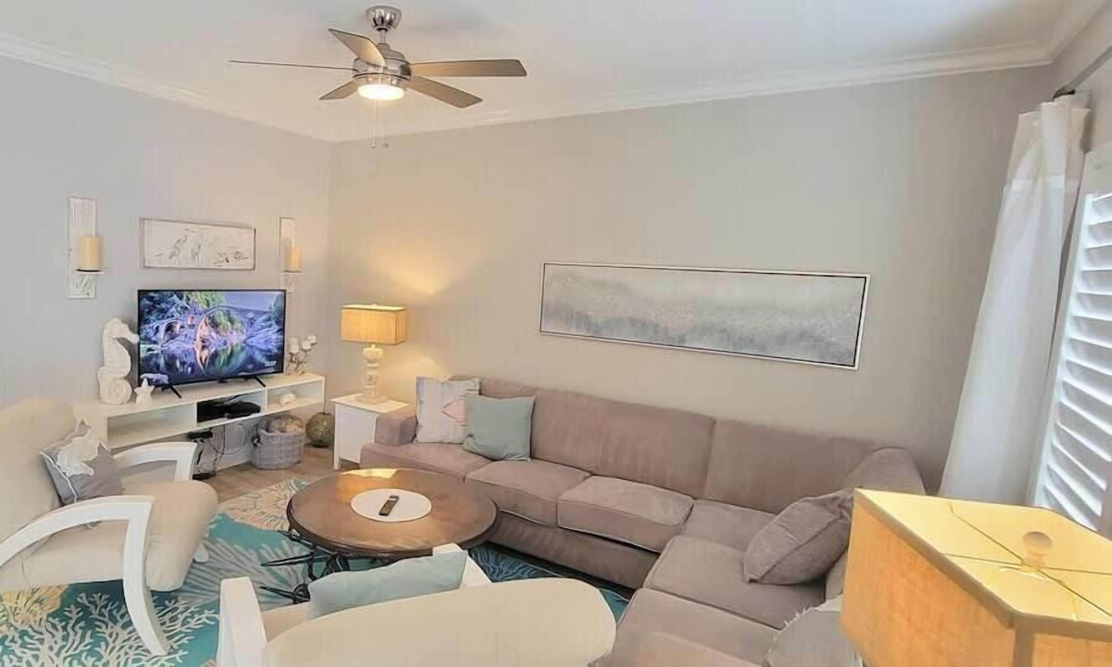 Un salon comprenant un canapé, des fauteuils, une table basse, un téléviseur, un ventilateur de plafond et un tableau accroché au mur.